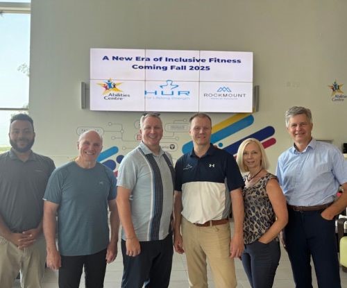 A  group of six people standing side by side in front of a digital sign that reads “A New Era of Inclusive Fitness – Coming Fall 2025” with the Abilities Centre, HUR, and Rockmount logos displayed.