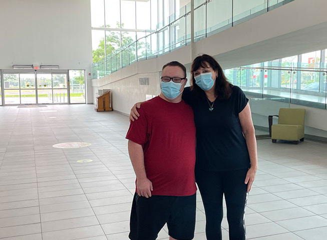Two Abilities Centre members stand wearing masks near the ramp in Abilities Centre Atrium on a sunny day. 