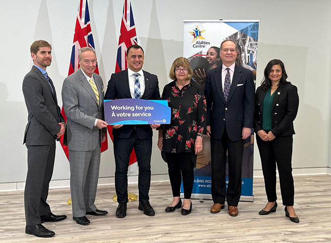 Abilities Centre senior leadership and members of the Government of Ontario smile before an Abilities Centre poster. From left to right the photo features:    Ross Ste-Croix, Abilities Centre COO; Lor