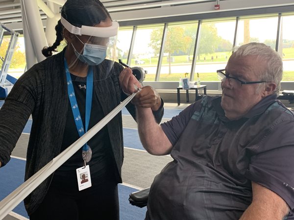 Abilities Centre trainer Andrea hold Dave's right hand while stretching it with a resistance band against a railing in the Fieldhouse