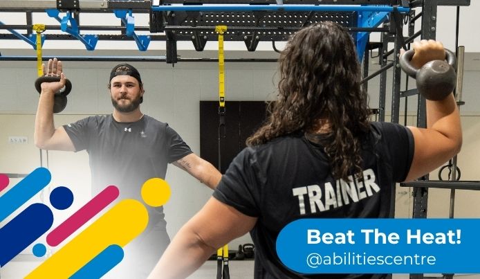 A personal trainer supports a client lifting a kettlebell inside the Fieldhouse at Abilities Centre. The text reads: “Beat The Heat! @abilitiescentre