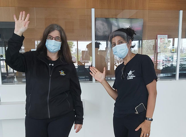 Two Abilities Centre staff members wearing black, blue masks and a face shield wave happily at the camera