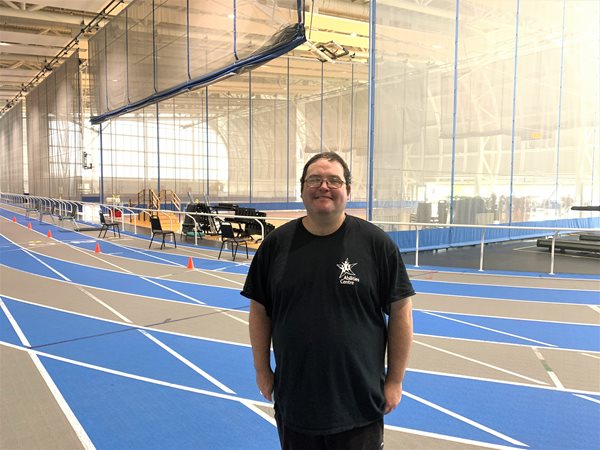 Adam stands in the Fieldhouse on the tracks smiling at the camera. He is wearing a black Abilities Centre shirt. 