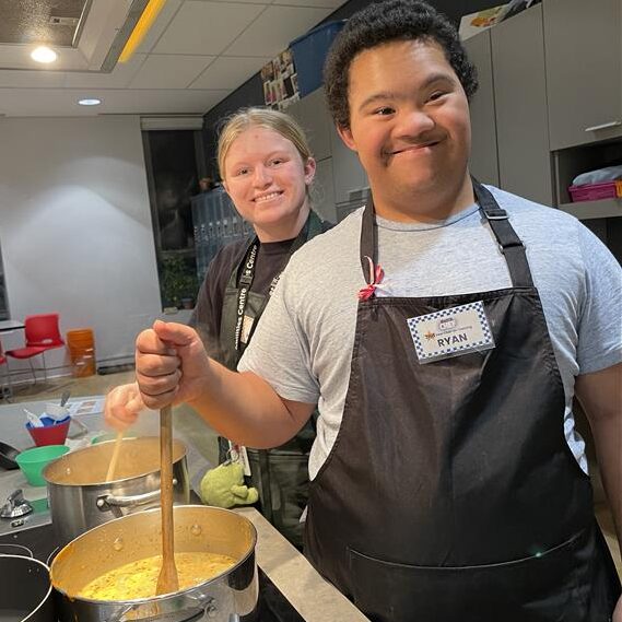A staff member and participant pose for a photo as they stir a sauce on the stove.