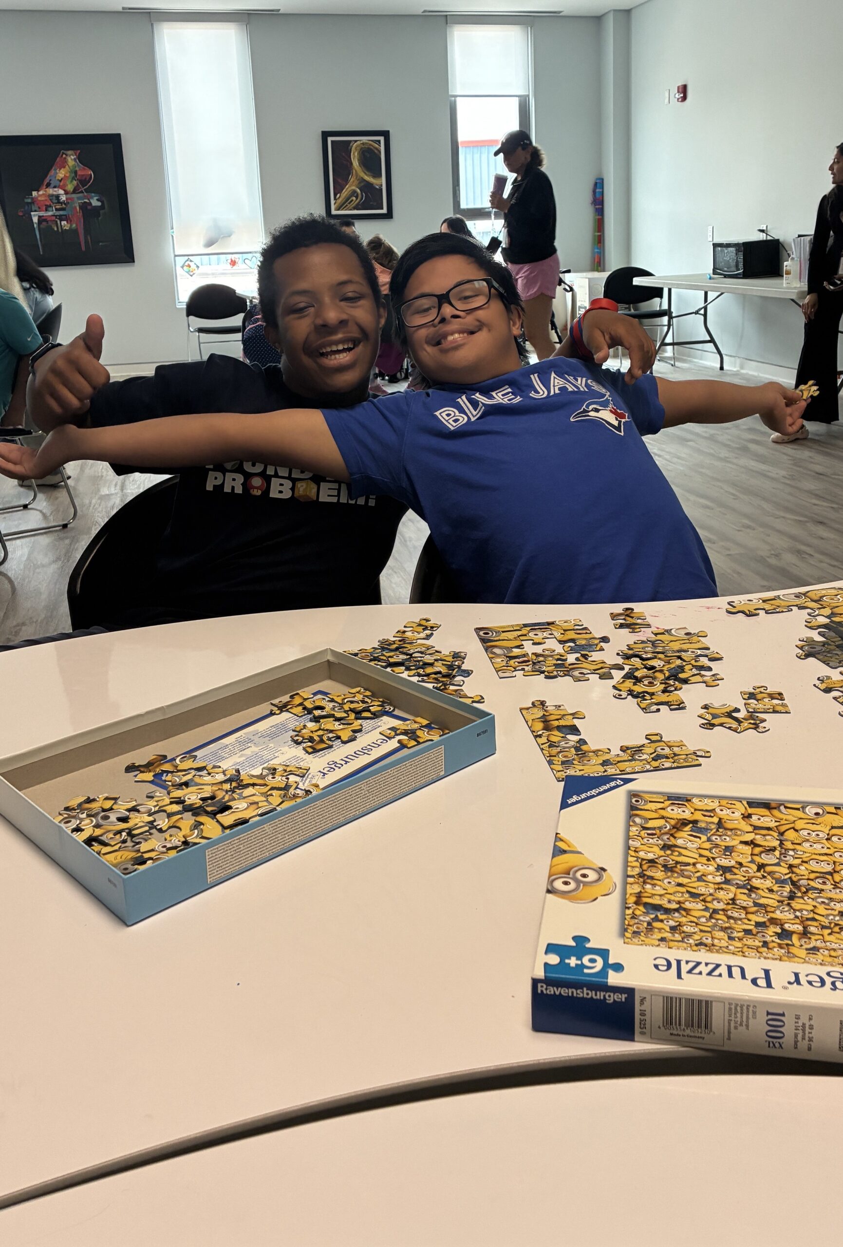 Two T R I S participants smile and pose with their arms wise open as they show off their in-progess Minions puzzle