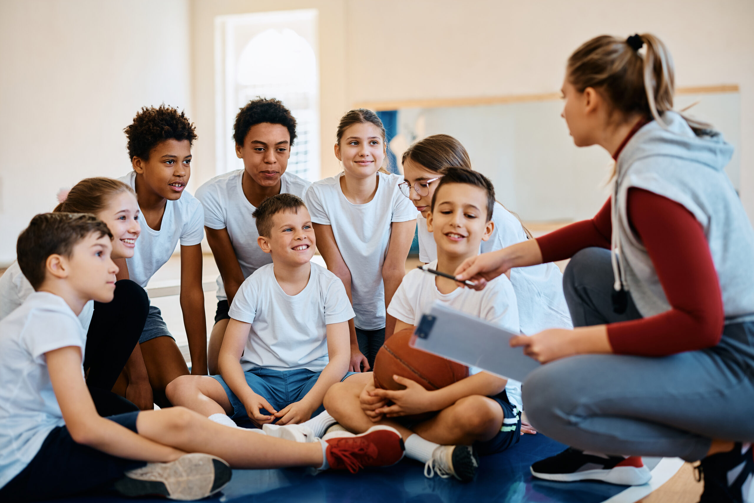 Happy elementary students listening basketball coach during physical education class at school gym.