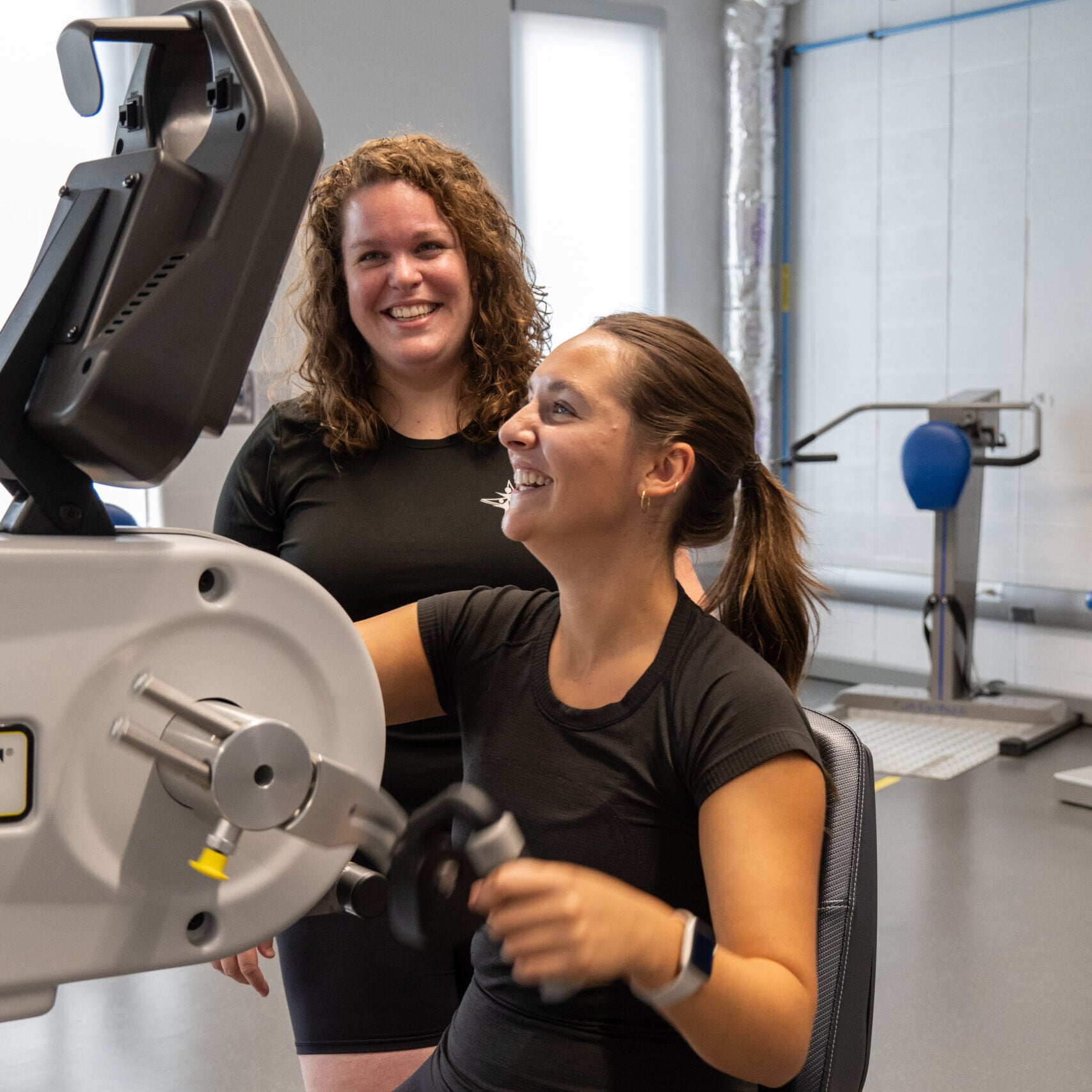 A personal trainer and her client smile as the client works out on an exercise machine.
