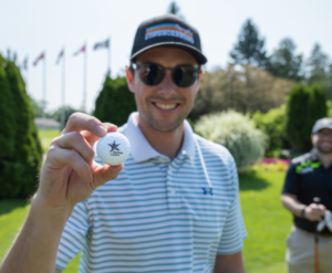 A man smiles while holding up a golf ball with the Abilities Centre logo on it.